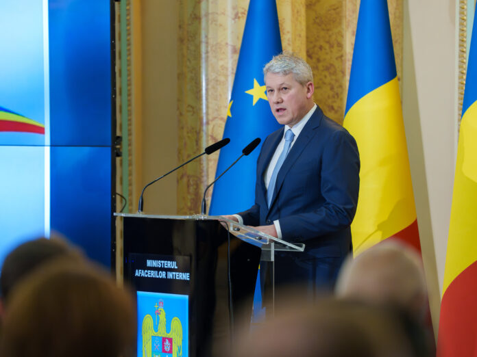 Cristian Predoiu, ministrul Afacerilor Interne, sustine o declaratie de presa la pupitrul MAI. Costum bleumarin, cravata bleu. Fundal cu steaguri Romania si UE, stema MAI pe pupitru. Atmosfera oficial