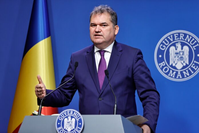 Cseke Attila-Zoltán susține declarații la pupitrul Guvernului, gesticulează cu mâna dreaptă. Steag românesc, fundal albastru cu siglă. Costum bleumarin, cravată mov-violet. Păr grizonat.