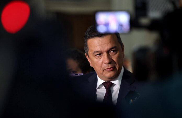 Sorin Grindeanu ofera declaratii presei intr-un spatiu interior intunecat. Costum bleumarin inchis cu cravata visinie. Privire concentrata spre jurnalisti, lumini de camere TV vizibile in fundal. Atmo