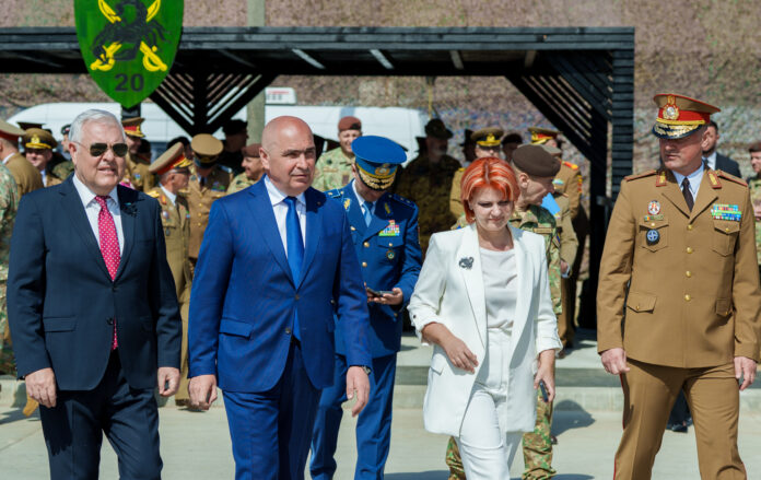 Ilie Bolojan si Lia Olguta Vasilescu la o baza militara, insotiti de oficiali militari in uniforma. Bolojan in costum bleumarin, Vasilescu in taior alb. Militari cu berete si uniforme de ceremonie. In
