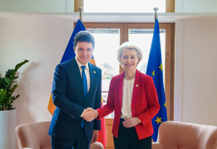 Nicusor Dan si Ursula von der Leyen, presedinta Comisiei Europene, dau mana intr-un salon oficial. Von der Leyen in sacou rosu, Nicusor Dan in costum bleumarin inchis. Steaguri UE si romanesc in funda