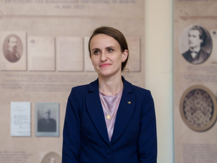 Oana Toiu, politician, portret in cadru institutional. Poarta sacou bleumarin peste bluza lila, insigna tricolora. Fundal cu panouri istorice despre reprezentarea romaneasca la Berlin. Expresie calma,