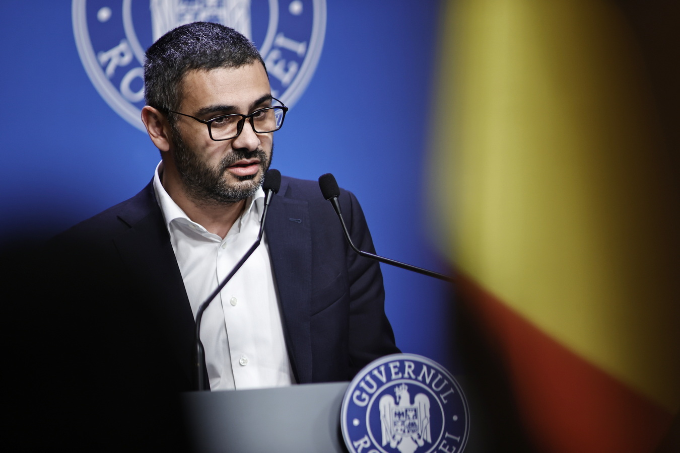 Transparentizarea salariilor: directiva UE, implementată la termen în consens cu sindicatele Florin Manole, politician roman, sustine declaratii la pupitrul Guvernului Romaniei. Barbat cu ochelari si barba, costum bleumarin inchis, camasa alba fara cravata. Fundal albastru cu stema Guvernului