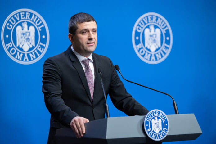 Radu Miruta, politician roman, sustine declaratii la pupitrul Guvernului Romaniei. Poarta costum bleumarin inchis cu cravata mov, pe fundal albastru cu stema Guvernului Romaniei. Microfoane pe pupitru