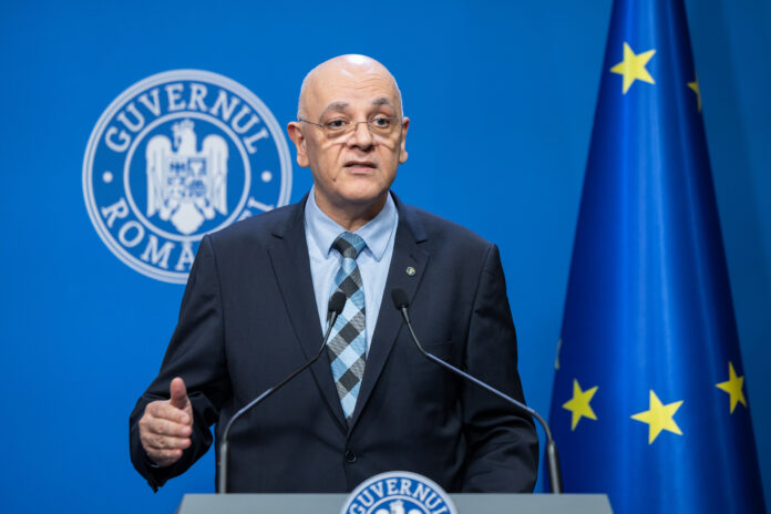 Raed Arafat, secretar de stat DSU, sustine declaratii la pupitrul Guvernului Romaniei. Barbat chel cu ochelari, costum bleumarin, camasa bleu, cravata cu model geometric. Steag UE si stema Guvernului