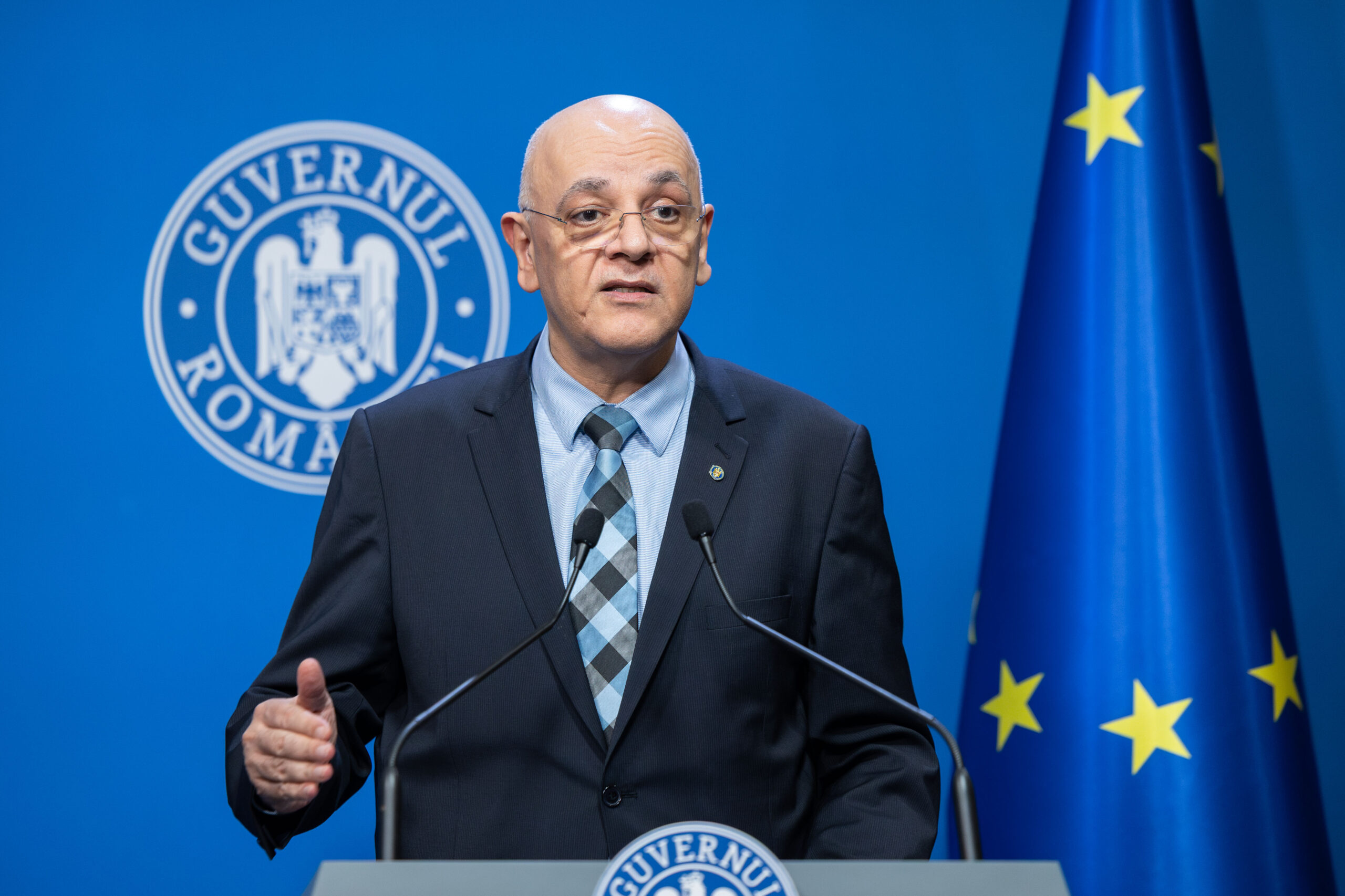 Arafat: Infirm categoric existenta unor angajari fictive la Departamentul pentru Situatii de Urgenta Raed Arafat, secretar de stat DSU, sustine declaratii la pupitrul Guvernului Romaniei. Barbat chel cu ochelari, costum bleumarin, camasa bleu, cravata cu model geometric. Steag UE si stema Guvernului