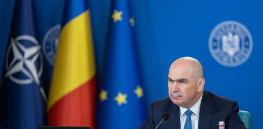 SURSE Criza carburanților: Măsurile Guvernului – până la 30 iunie Ilie Bolojan la ședință de guvern, cu laptop deschis, steaguri NATO, România și UE în fundal, siglă Guvern, cadru de lucru