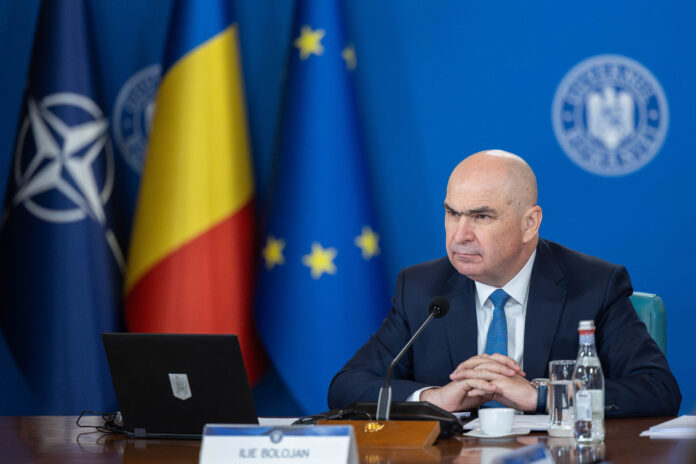 Ilie Bolojan la ședință de guvern, cu laptop deschis, steaguri NATO, România și UE în fundal, siglă Guvern, cadru de lucru