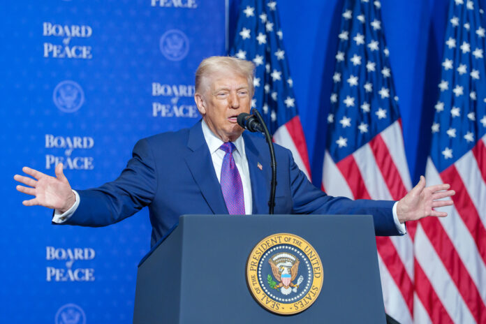 Donald Trump susținând un discurs la Board of Peace, cu brațele deschise la pupitrul prezidențial, steaguri americane în fundal