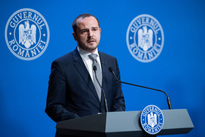 Alexandru Rogobete la pupitrul Guvernului României, costum bleumarin cu cravată gri, siglă Guvernul României, cadru oficial