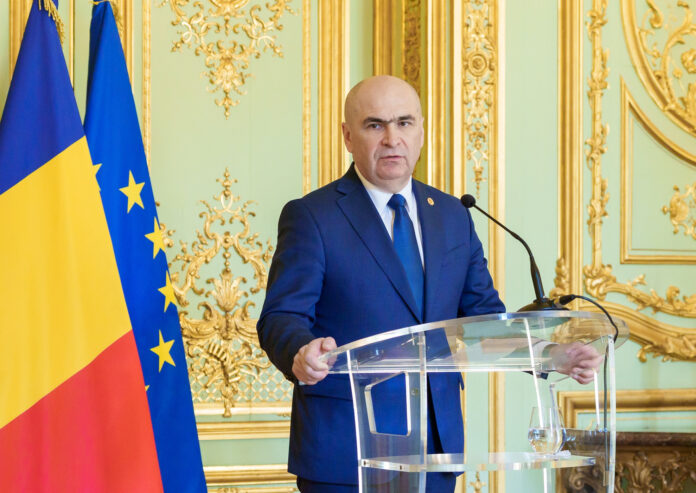 Ilie Bolojan la ședință de guvern, cu laptop deschis, steaguri NATO, România și UE în fundal, siglă Guvern, cadru de lucru