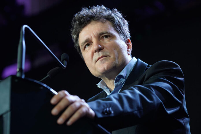 Nicusor Dan, presedintele Romaniei, la pupitru transparent cu decor auriu elegant. Costum bleumarin, cravata albastra inchisa. Steaguri romanesti si UE in fundal. Cadru formal, sala de protocol.