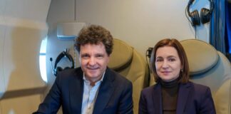 Nicușor Dan și Maia Sandu – în același avion către reuniunea Comunității Politice Europene de la Erevan Nicusor Dan sustine un discurs la pupitrul Ambasadei Romaniei (inscriptia 'Ambasada Romaniei'). Costum bleumarin, cravata albastra, gest larg cu mana. Steag romanesc si american in fundal. Fundal alba